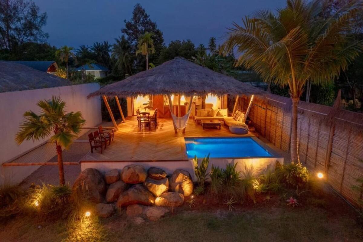 A thatched-roof outdoor lounge area with a pool, surrounded by rocks and palm trees.
