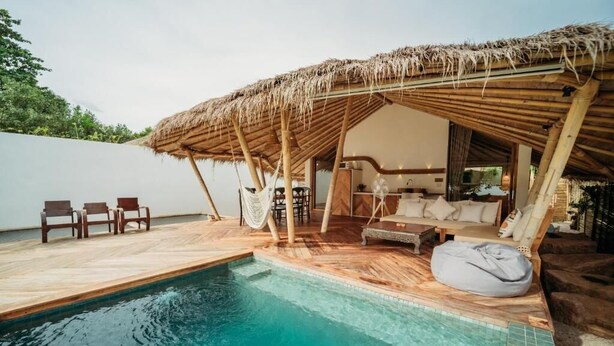 Un espace piscine doté d’un coin salon avec toit de chaume, d’une terrasse en bois et de chaises longues.