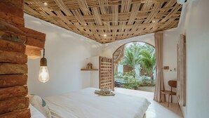 Une chambre à coucher avec un lit, un luminaire suspendu, une tête de lit en bois, une fenêtre donnant sur des palmiers et une porte menant à l’extérieur.