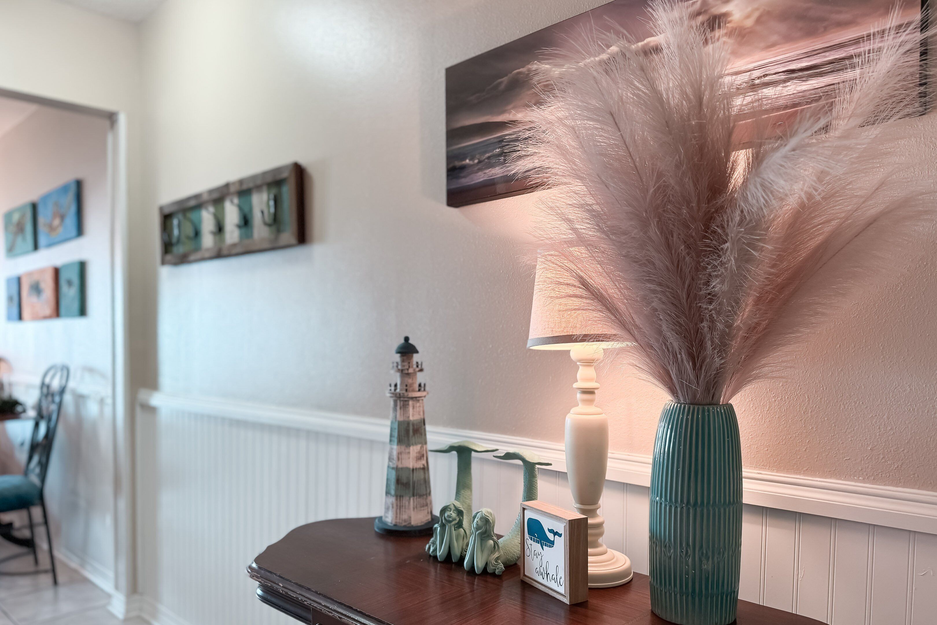 Una mesa de madera con elementos decorativos, incluyendo un faro, una imagen enmarco de una ballena y un jarrón con plantas secas.