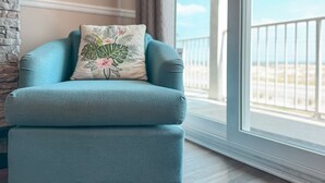 Un fauteuil turquoise agrémenté d’un coussin à motifs floraux, placé à côté d’une cheminée en pierre, et offrant une vue sur la plage par le biais d’une grande fenêtre.