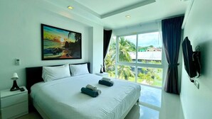 A hotel room with a large bed, a painting of a beach scene, a TV, and a window with curtains. - 6-17 - 2 Bdr Condo with Balcony and shared Pool (Tambon Patong)