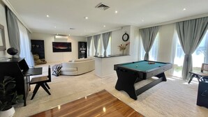 A modern living room with a pool table, a sofa, a dining area, and a large window with curtains. - Brighton Blissful | Family Poolside Vila Pool Spa Sauna (Brighton East)