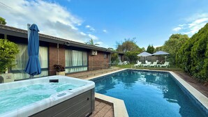 A swimming pool with a hot tub, surrounded by a brick house, a covered patio, and a lawn with lounge chairs. - Brighton Blissful | Family Poolside Vila Pool Spa Sauna (Brighton East)