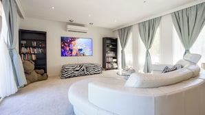 A modern living room with a white sofa, a zebra-patterned ottoman, and two bookshelves filled with books. - Brighton Blissful | Family Poolside Vila Pool Spa Sauna (Brighton East)