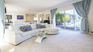 A spacious living room with a white sofa, a glass coffee table, and a large window with curtains. - Brighton Blissful | Family Poolside Vila Pool Spa Sauna (Brighton East)