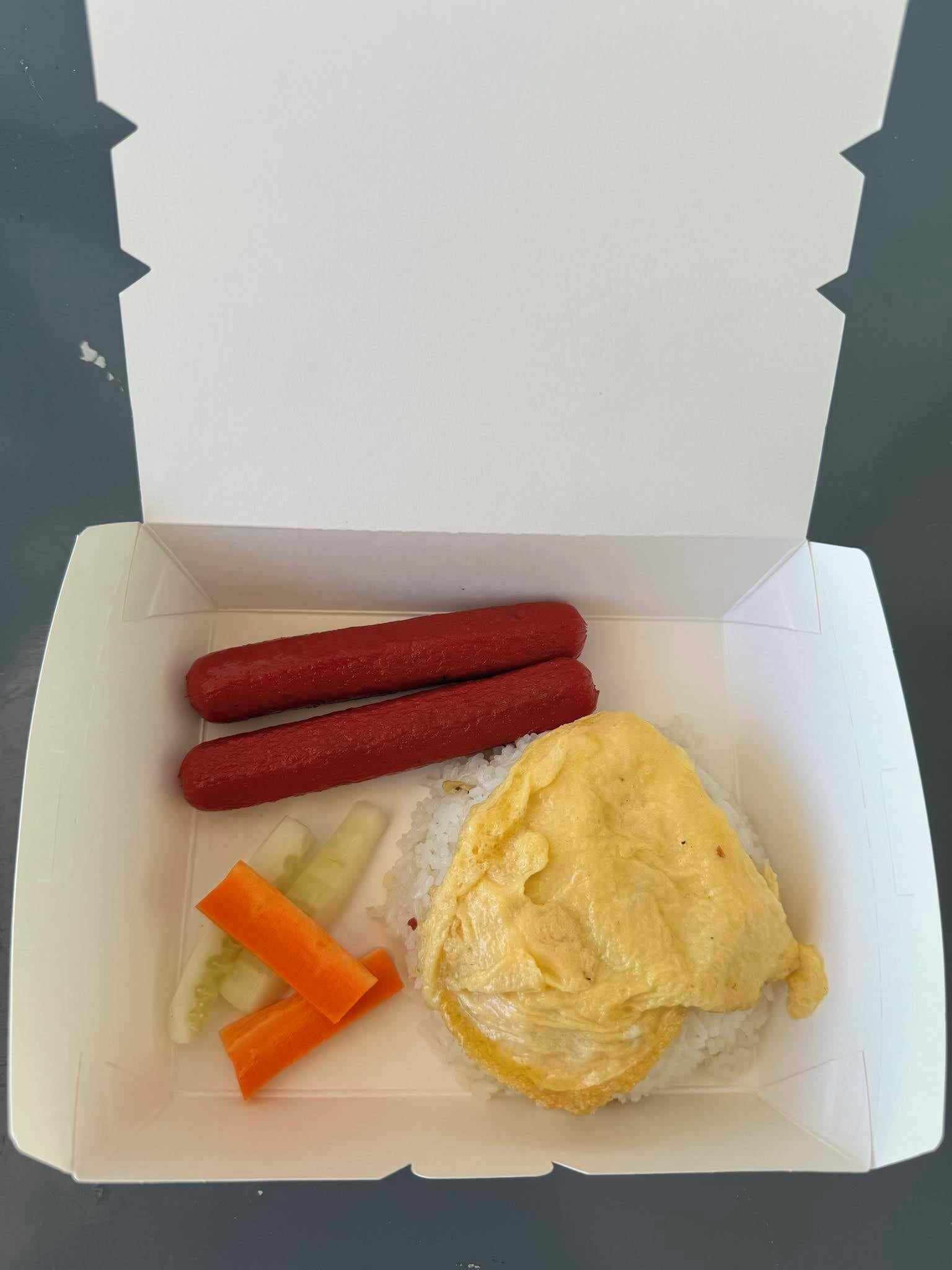Una caja para llevar abierta que contiene dos perritos calientes, un huevo frito, pepinillos y zanahorias.