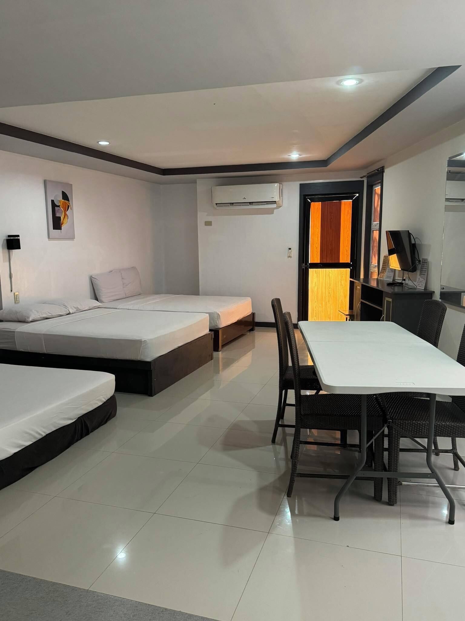 A modern hotel room with a large bed, a smaller bed, a dining table with chairs, an air conditioning unit, and a TV.