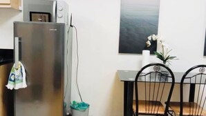 Ett kompakt kök med ett kylskåp, en mikrovågsugn och ett litet matbord med två stolar. Ovanför kylskåpet finns en hylla med krukväxter, och bredvid står en liten papperskorg.