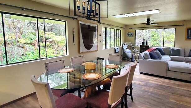 Un comedor con mesa y sillas de vidrio, un sofá y ventanas amplias con vistas a zonas verdes.