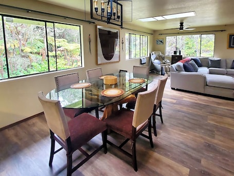 A dining area with a glass table and chairs, a sofa, and large windows with a view of greenery.