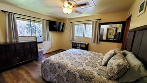 Un dormitorio con cama, ventilador de techo, cómoda, televisor y ventanas con cortinas.