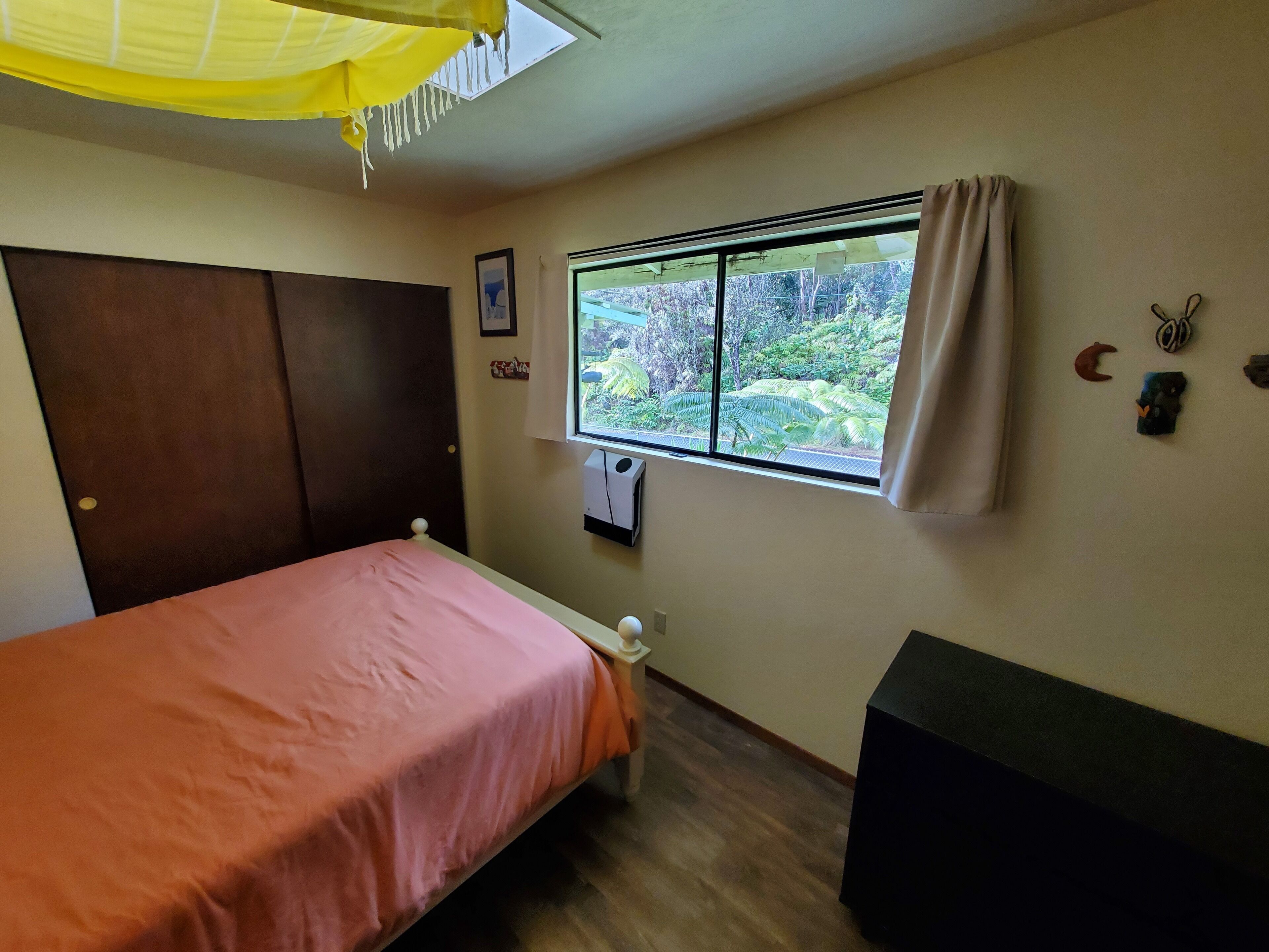 A bedroom with a bed, a window with a view of greenery, a ceiling fan, and a wall-mounted heater.