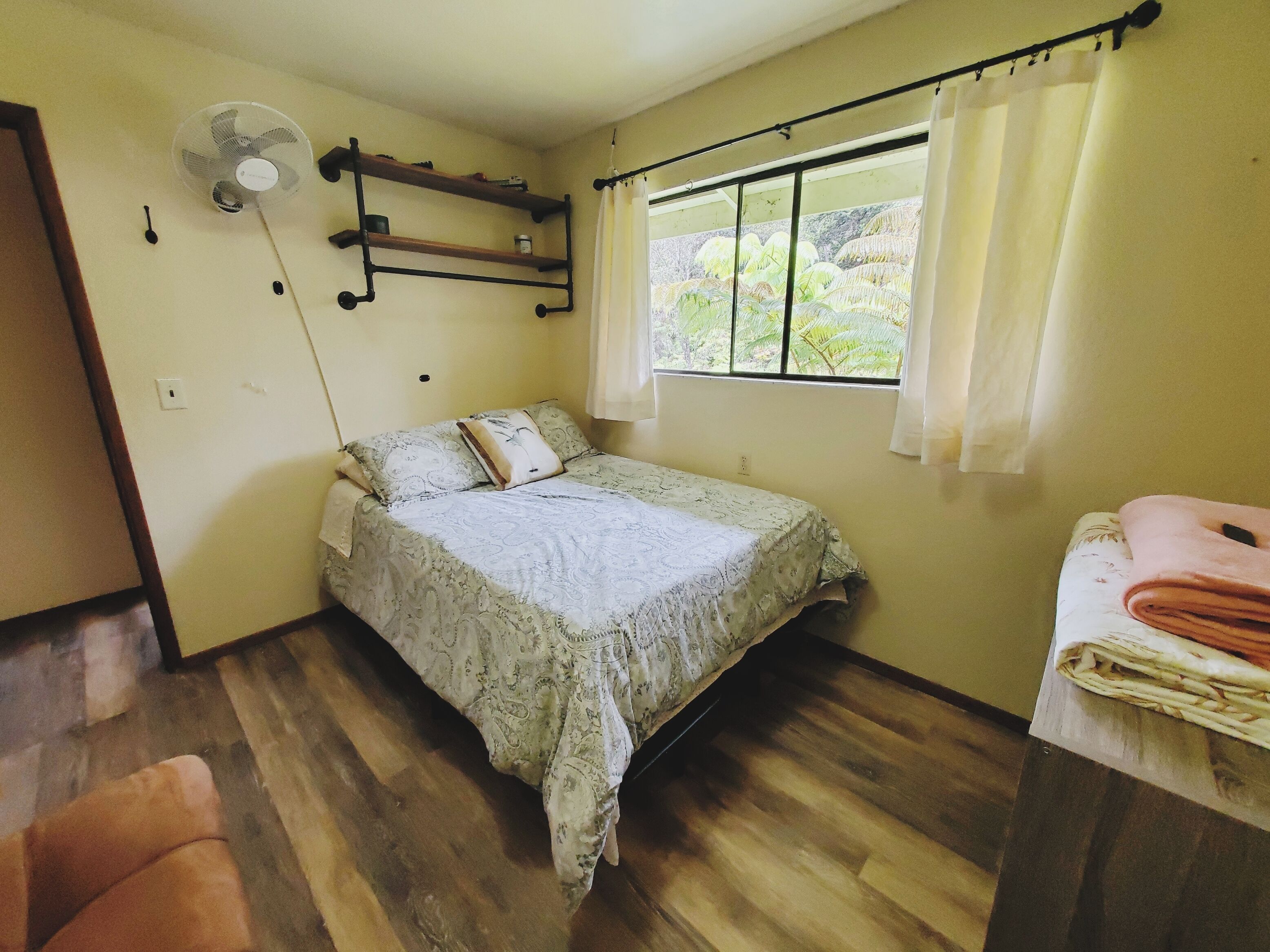 A small, cozy bedroom with a single bed, a bedside table with a folded towel, a standing fan, and a window with curtains.