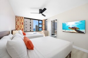 Quarto de hotel com duas camas, um ventilador de teto, uma janela grande com vista para a cidade e uma fotografia emoldurada de uma tartaruga marinha.