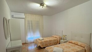 A room with two beds, a small table, a nightstand, an air conditioning unit, and a window with curtains. - The Beautiful Views II (Noalejo)