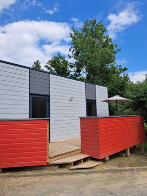 Ein modernes Gebäude mit grauer Fassade und roten Akzenten, das eine Holzterrasse und einen überdachten Außenbereich bietet.