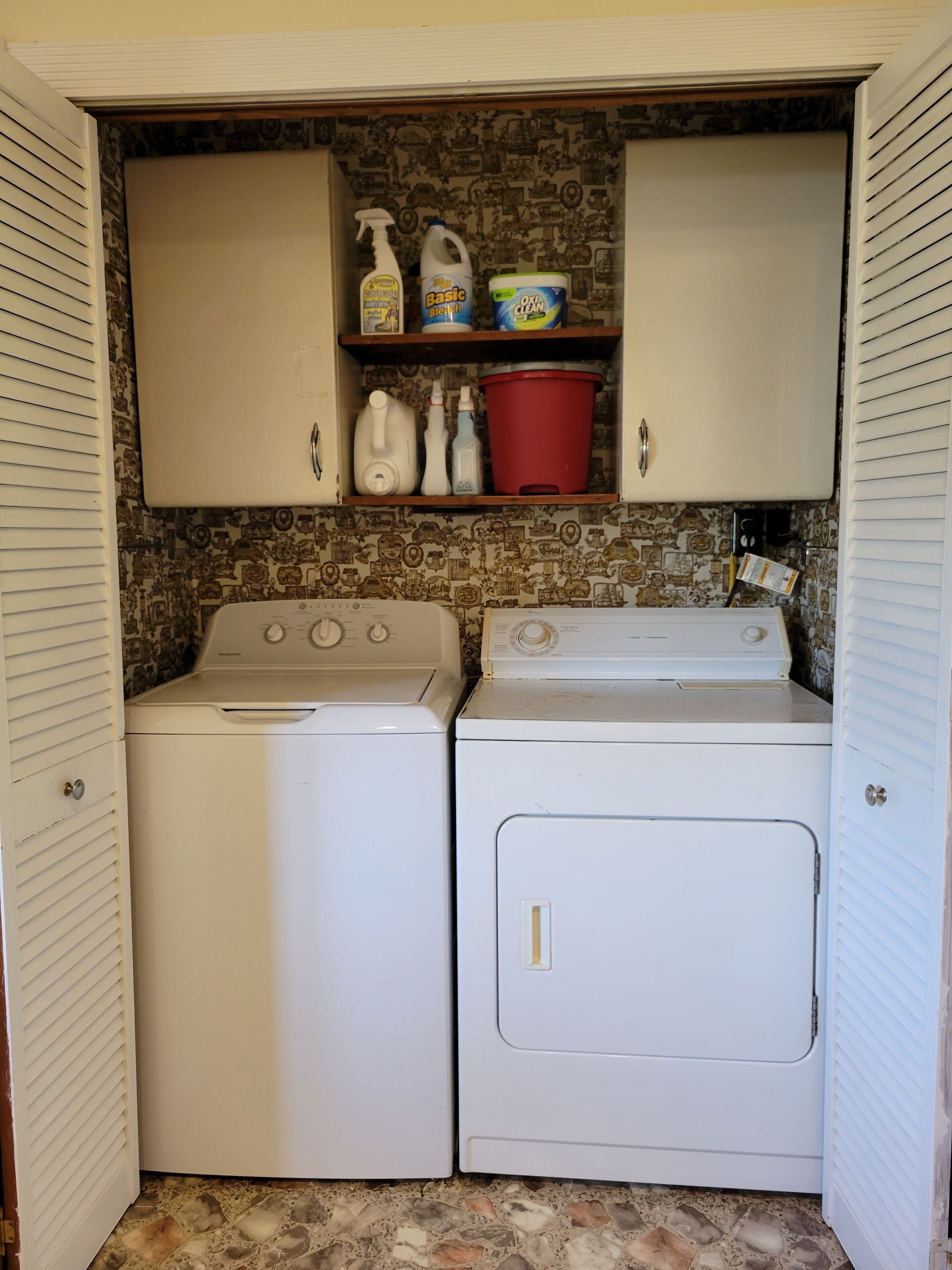 A laundry room with two white washing machines, a shelf with cleaning supplies, and a red bucket.
