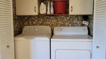 A laundry room with two white washing machines, a shelf with cleaning supplies, and a red bucket.
