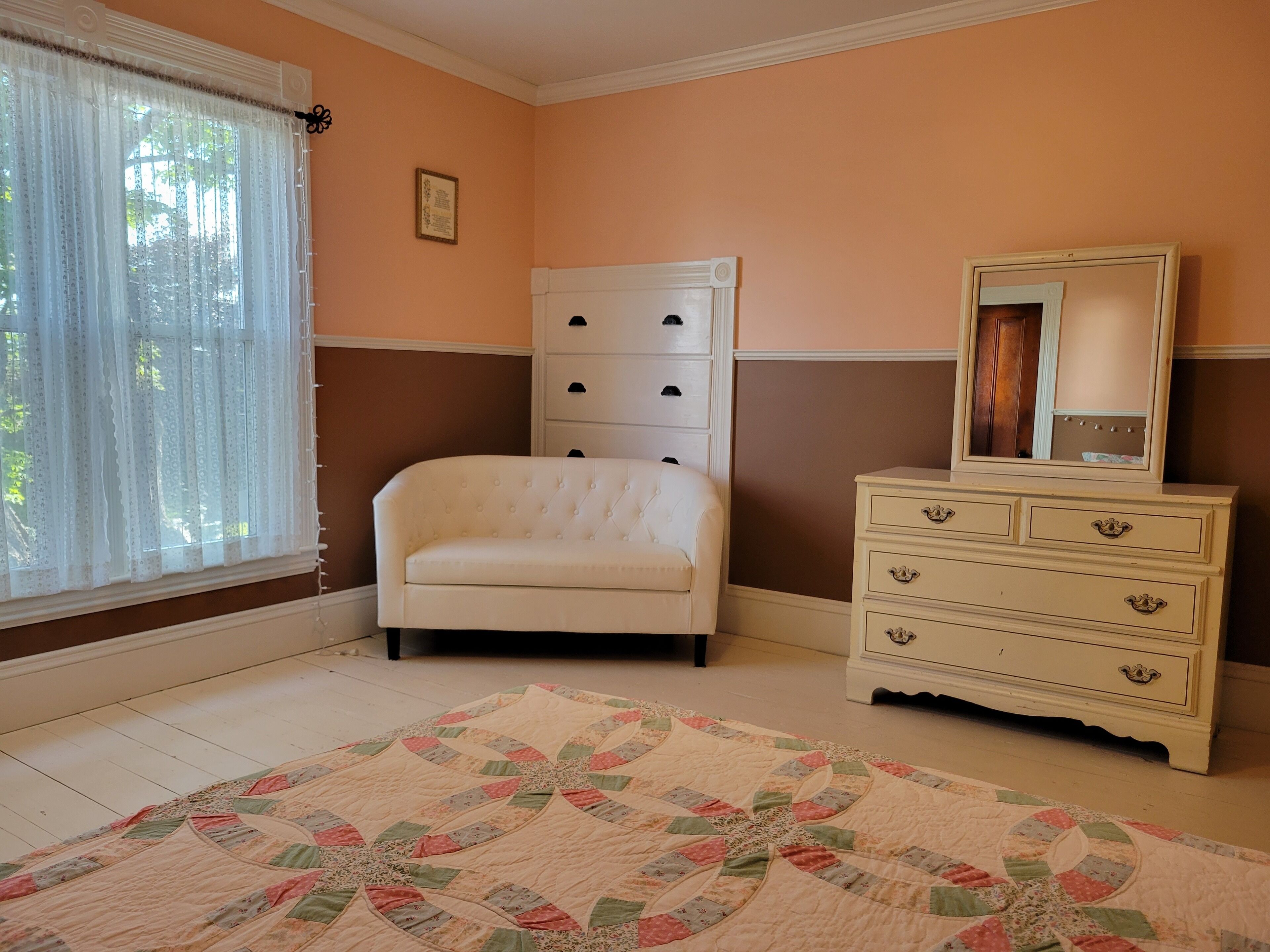 Un dormitorio con una silla blanca, un aparador con cajones y un colcha en el suelo.