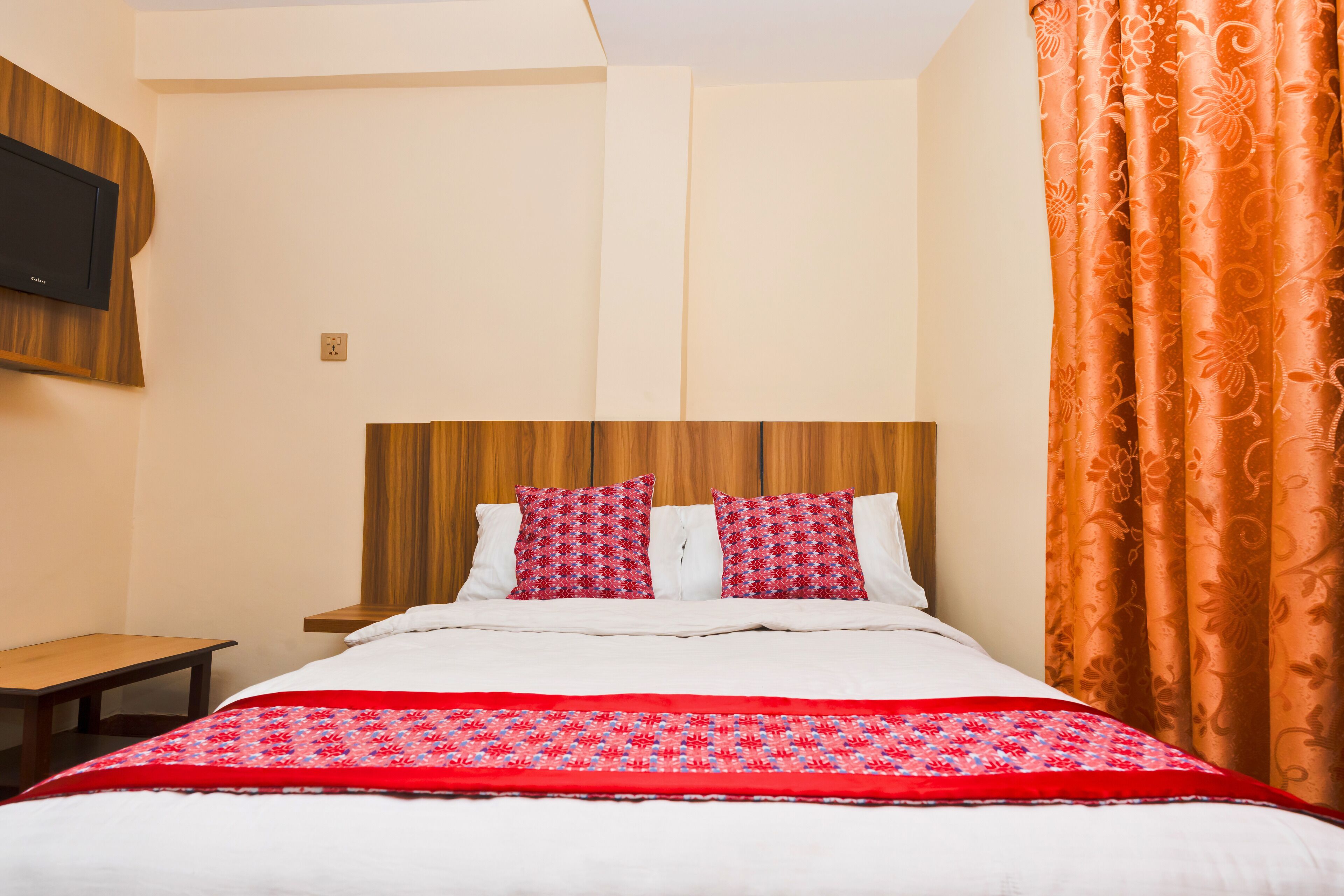 A hotel room with a wooden headboard, two red patterned pillows, a red and white bedspread, and an orange curtain.