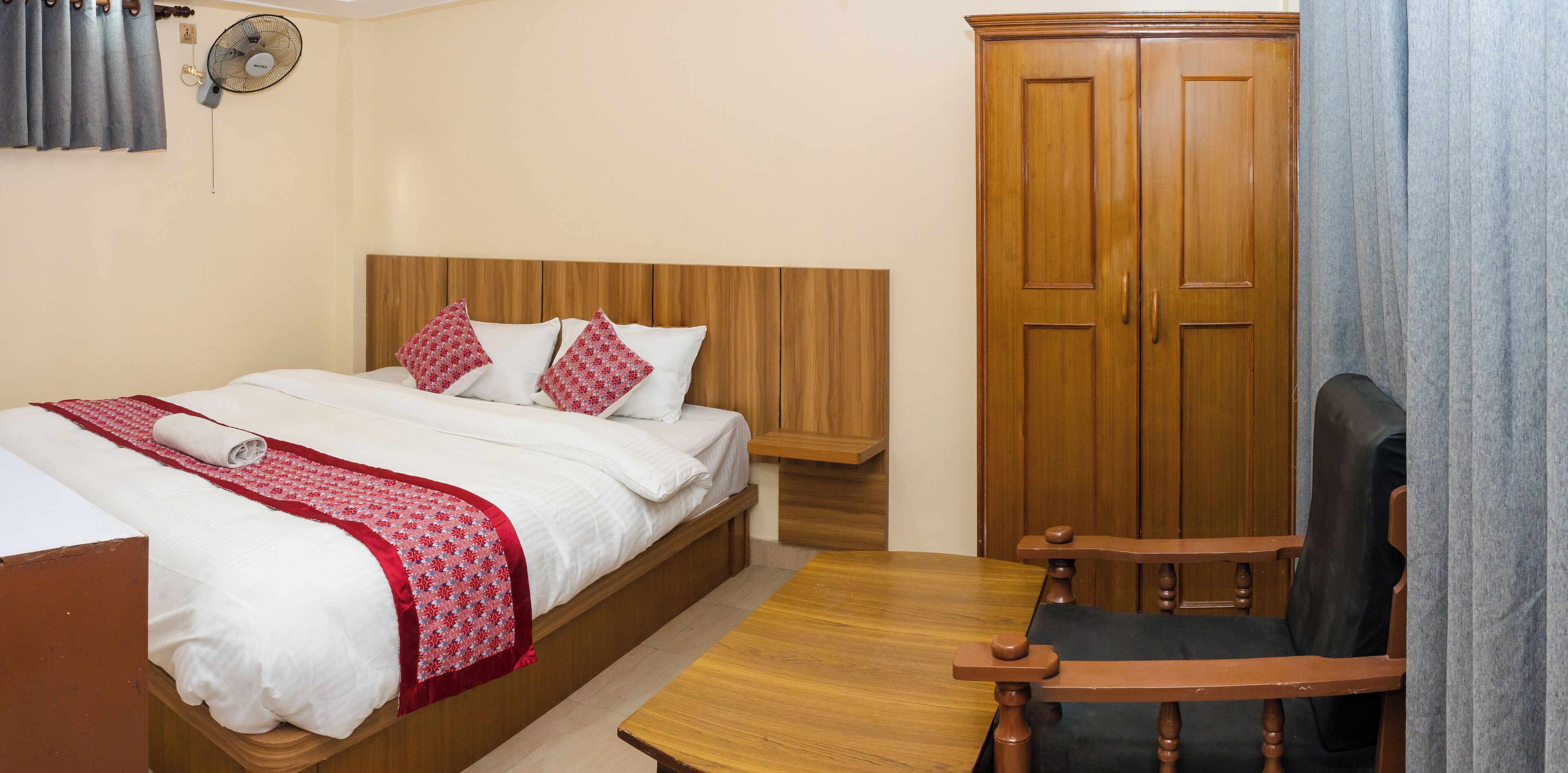 A hotel room with a bed, a wooden wardrobe, a wooden table, and a chair.