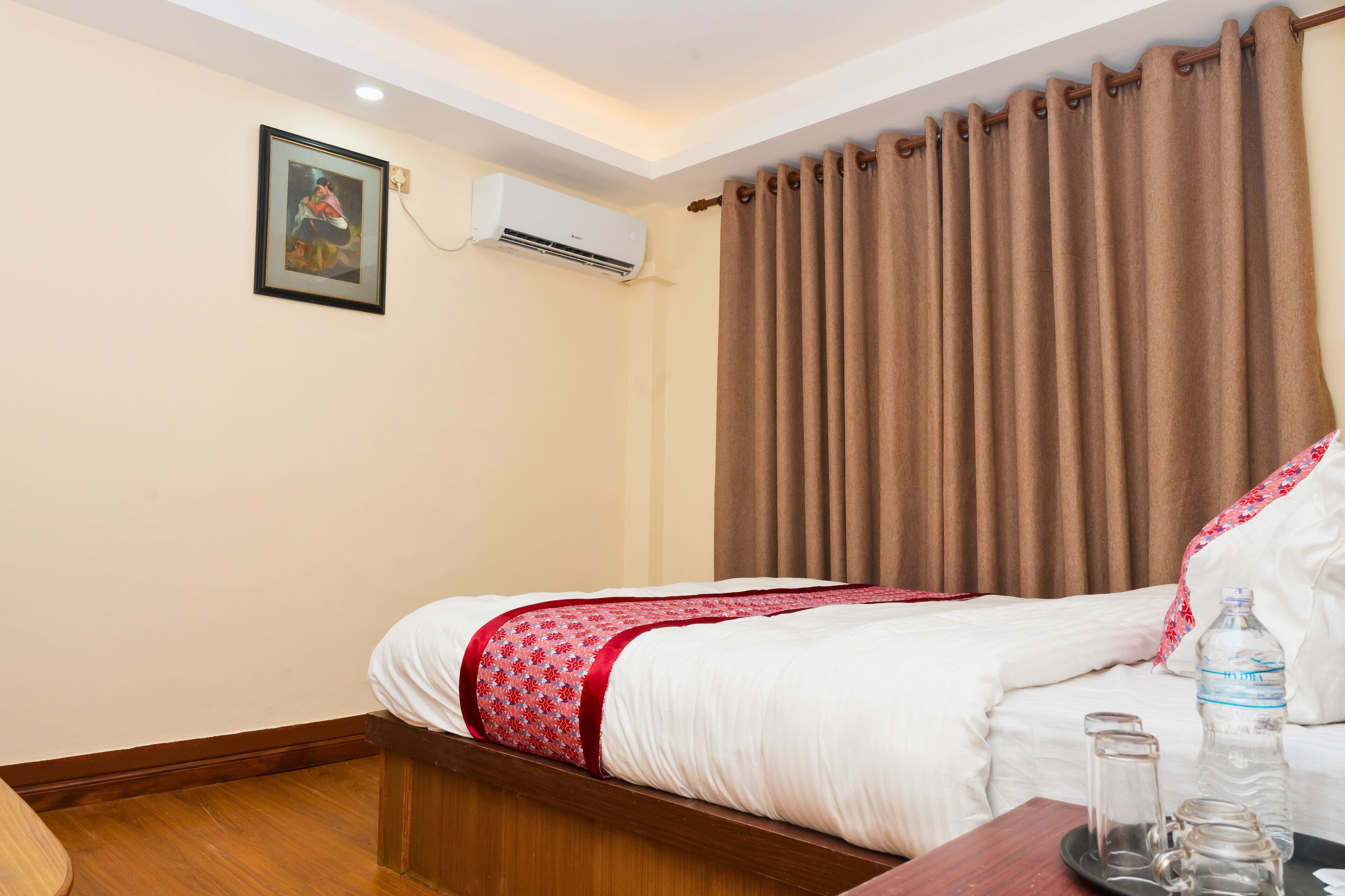 A hotel room with a bed, a bedside table with a bottle and glasses, a picture on the wall, and an air conditioning unit.