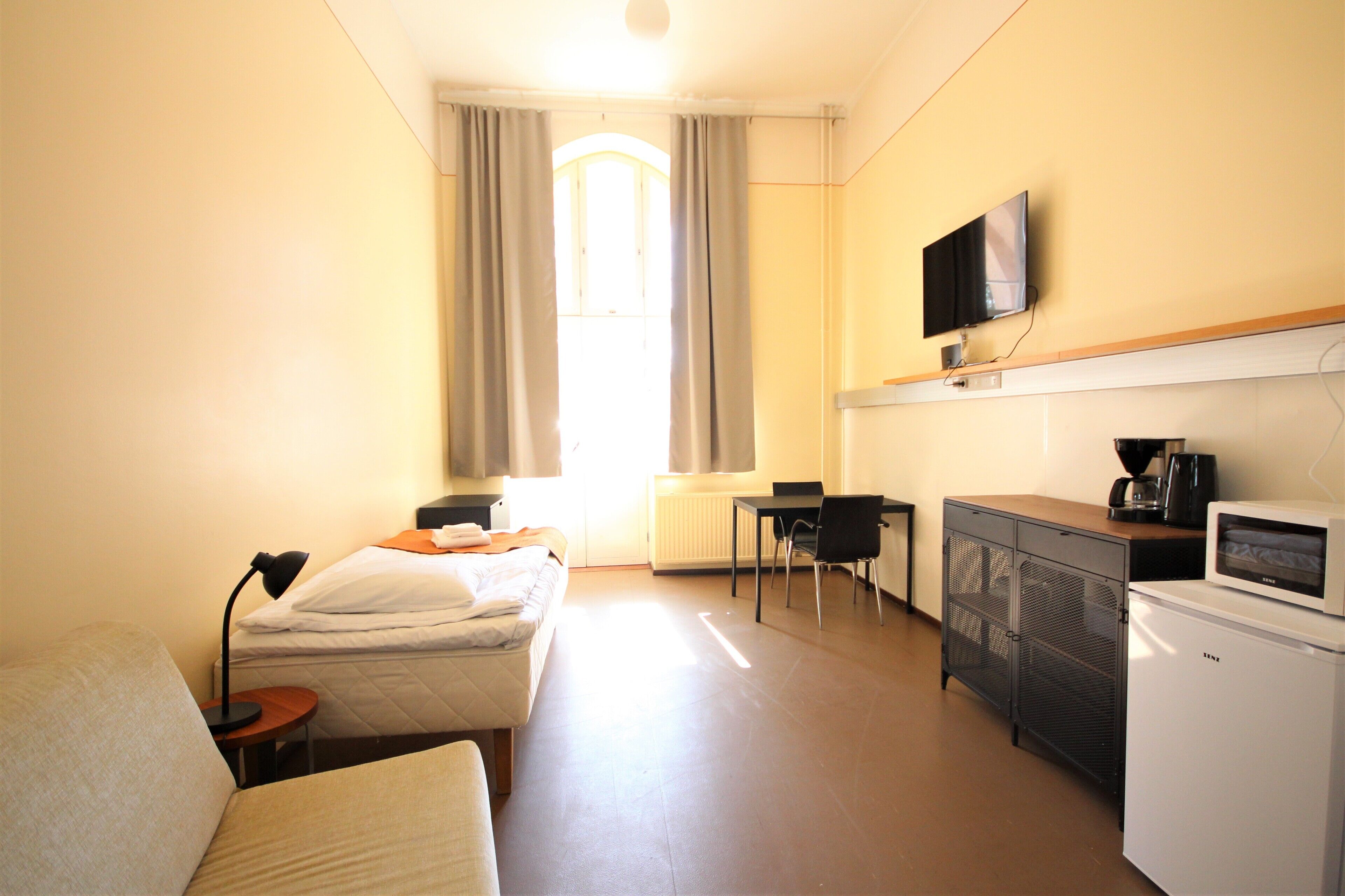 Hotellihuone, jossa on sänky, työpöytä, televisio, jääkaappi ja ikkuna, jossa on verhot.