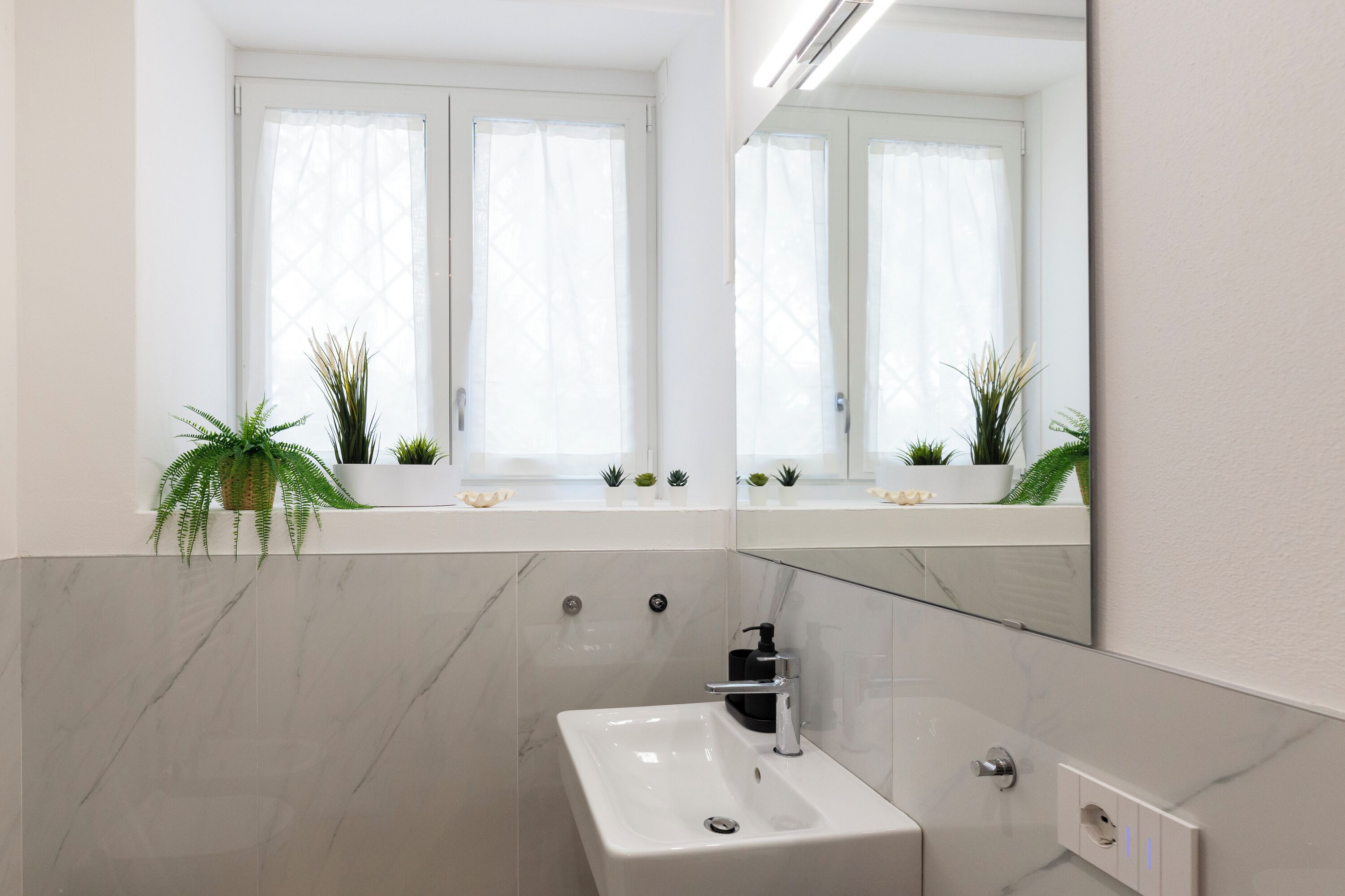 Un baño moderno con un lavamanos blanco, un espejo y una ventana con cortinas.