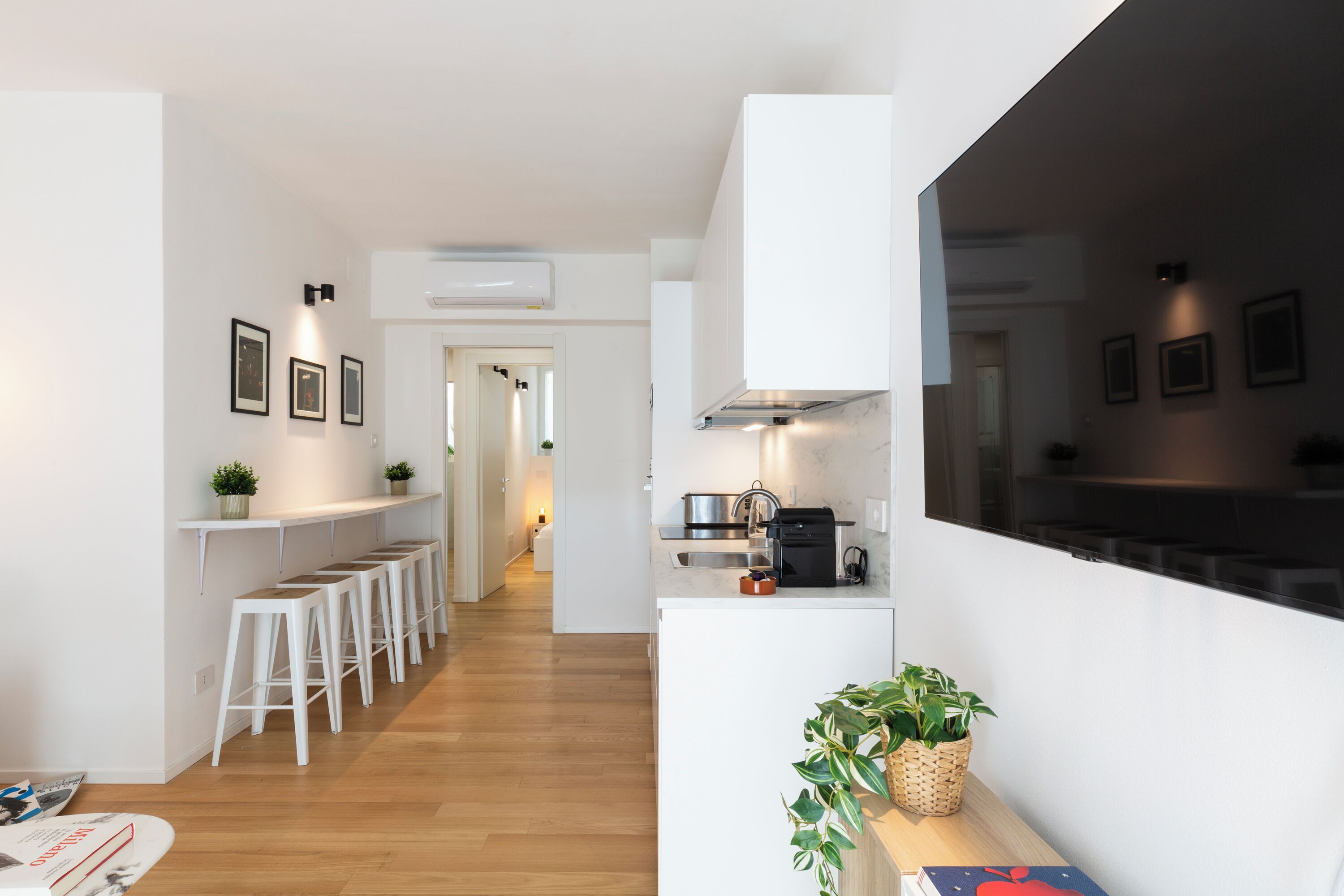 A modern kitchen with white cabinetry, a built-in oven, and a coffee machine. There are bar stools along the wall and a plant on the counter.