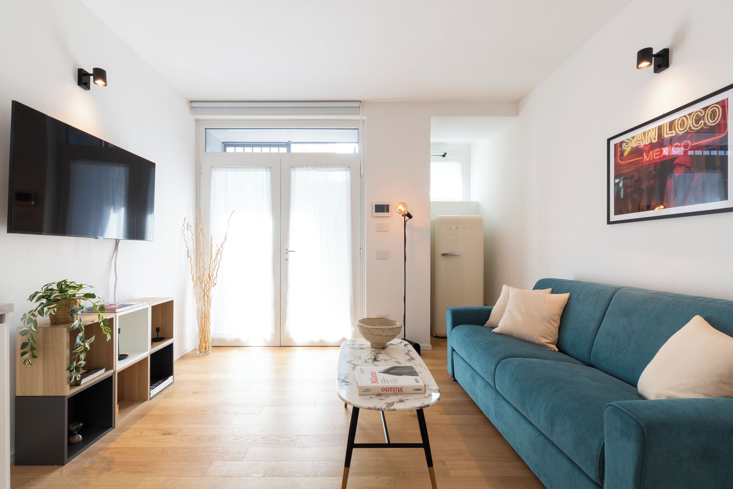 A modern living room with a teal sofa, a flat-screen TV mounted on the wall, and a framed picture of San Francisco.