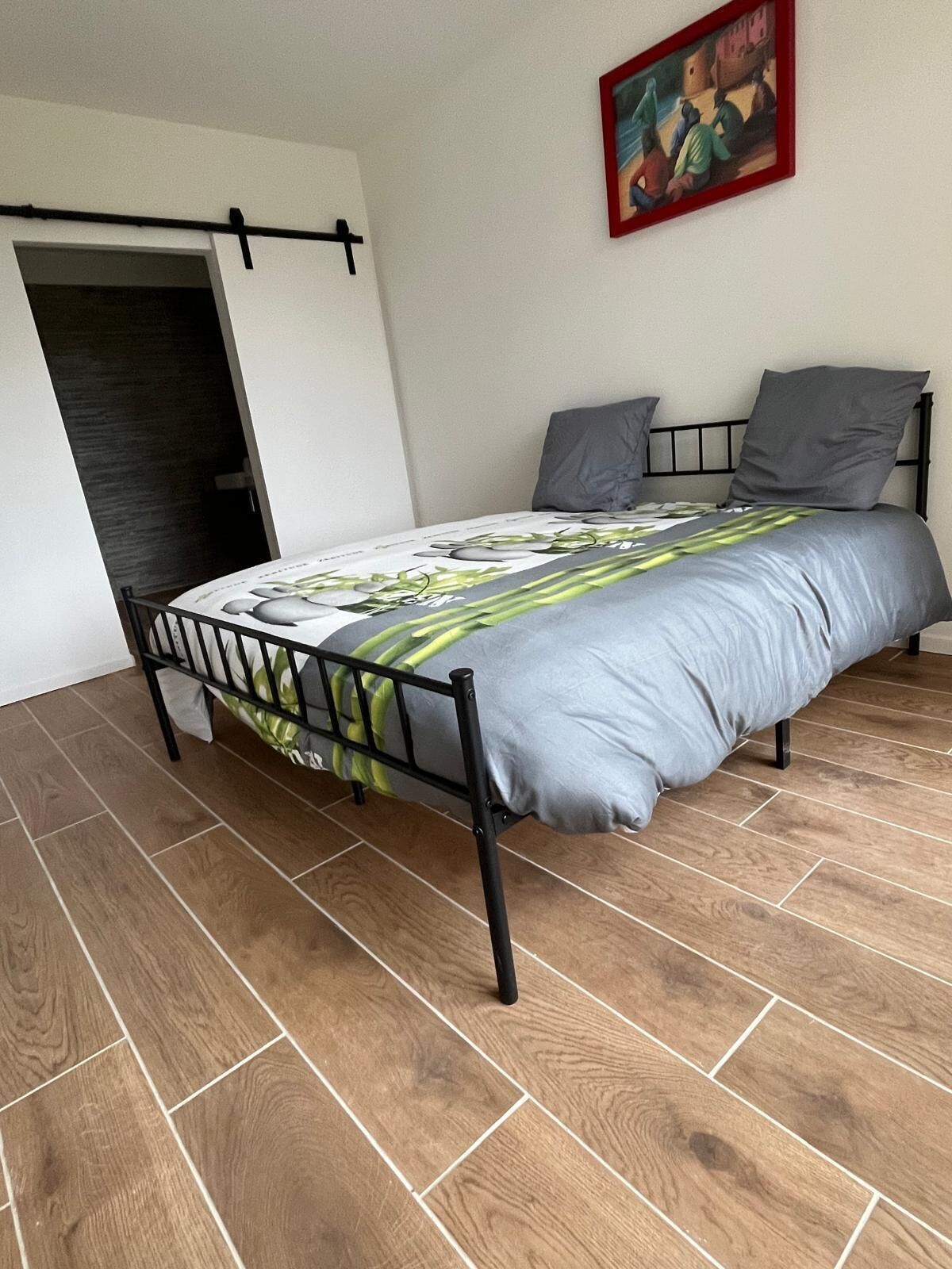 Une chambre à coucher avec un lit, du linge de lit gris, un plancher en bois, un tableau au mur et une porte donnant sur une autre pièce.