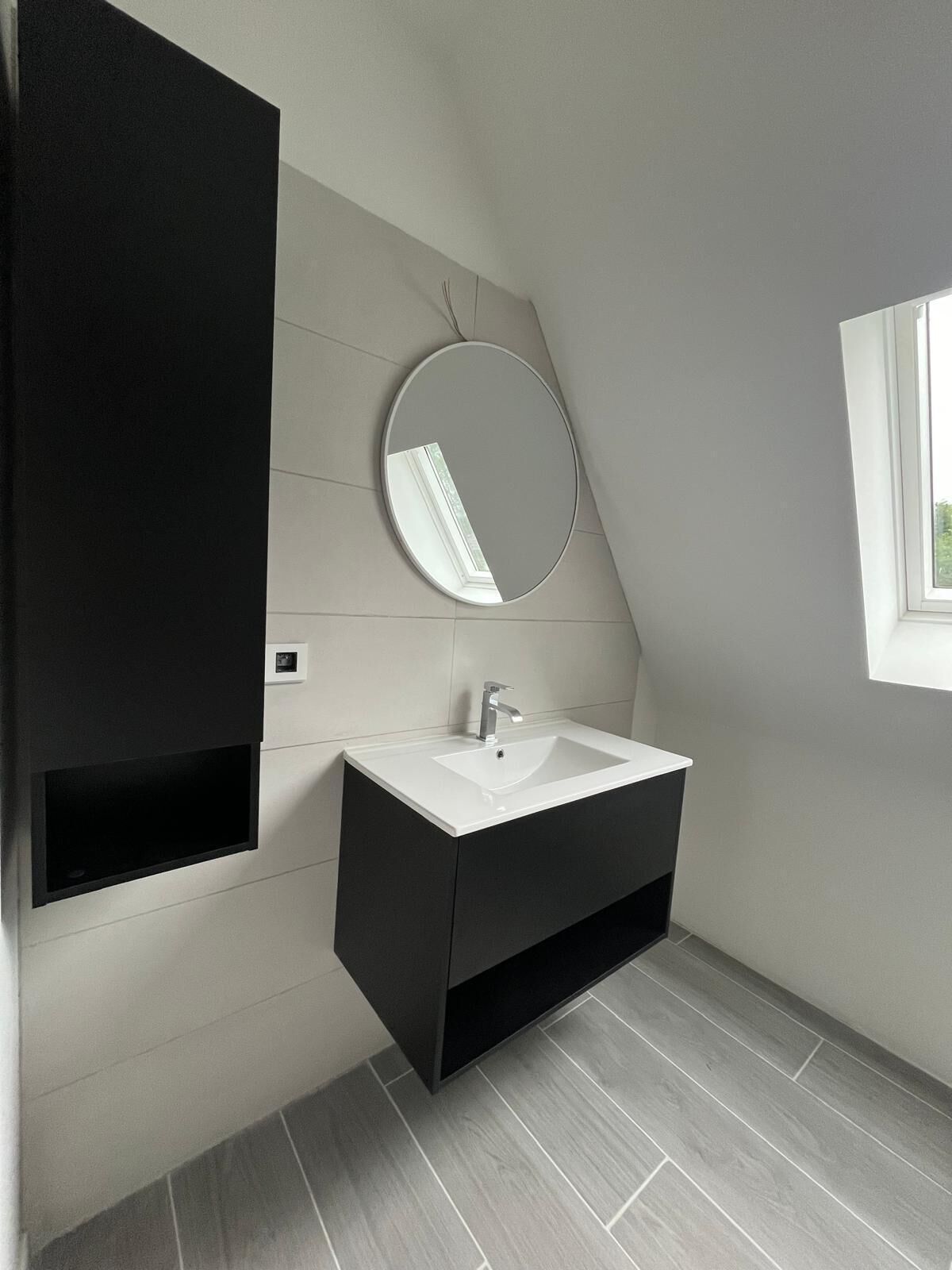 Een moderne badkamer met een witte wastafel, donkerd e badkamerkast en een ronde spiegel.