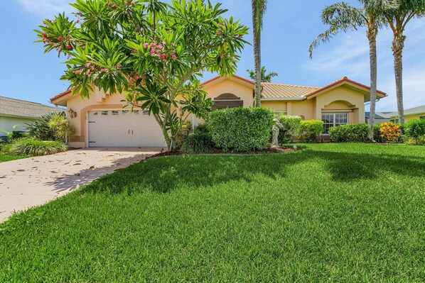 A residential house with a well-maintained lawn, a garage, and a large tree with pink flowers. - Gorgeous, Gulf Access canal, Heated Pool, Kayaks, Bicycles - Villa Mango - Roelens (Cape Coral)