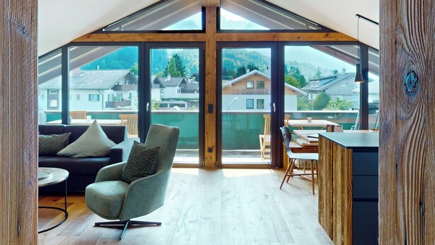 A spacious room with a high ceiling, large windows, a modern chair, a dining table, and a wooden cabinet.