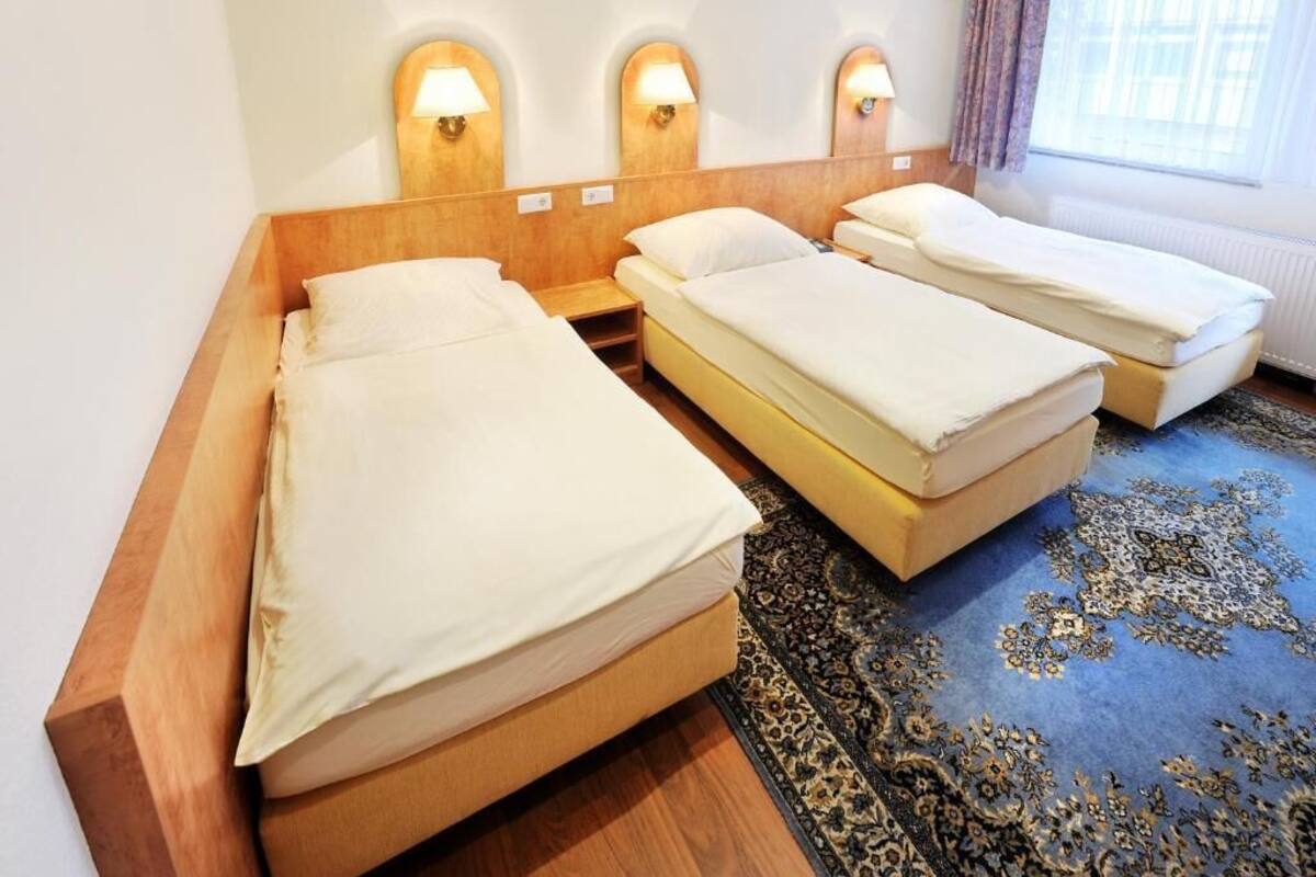 Quarto de hotel com três camas de solteiro, cabeceira de madeira, luminárias fixadas na parede e carpete estampado.