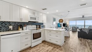 A modern kitchen with white cabinets, a microwave, and a dishwasher. There is a dining area with a gray sofa and a view of the balcony. - Oceanfront 3BR condo with pool, balcony, and beach access. (Fernandina Beach)