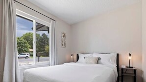 Un dormitorio con una cama grande, una mesita de noche con una lámpara y vistas a la calle por una ventana.