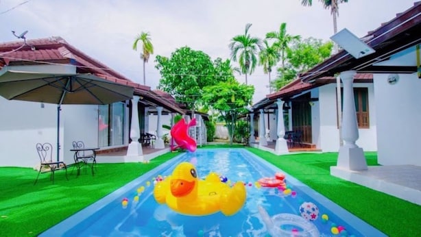 A swimming pool with inflatable toys, surrounded by white buildings with red roofs and green lawns. - Karnie Cheer Pool Villa & Resort Pattaya (Sattahip)