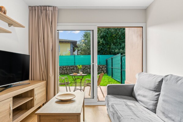 Un salon moderne avec un téléviseur à écran plat, un canapé gris, une table basse en bois et une porte-fenêtre coulissante donnant sur une terrasse avec une clôture verte et un petit jardin.