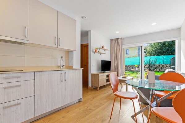 Una cocina moderna con armarios de madera clara, una zona de comedor con una mesa de vidrio y sillas naranjas, y un televisor al fondo.