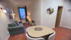 Een woonkamer met een groene bank, een houten eettafel met stoelen, een televisie en een raam met uitzicht op gebouwen.
