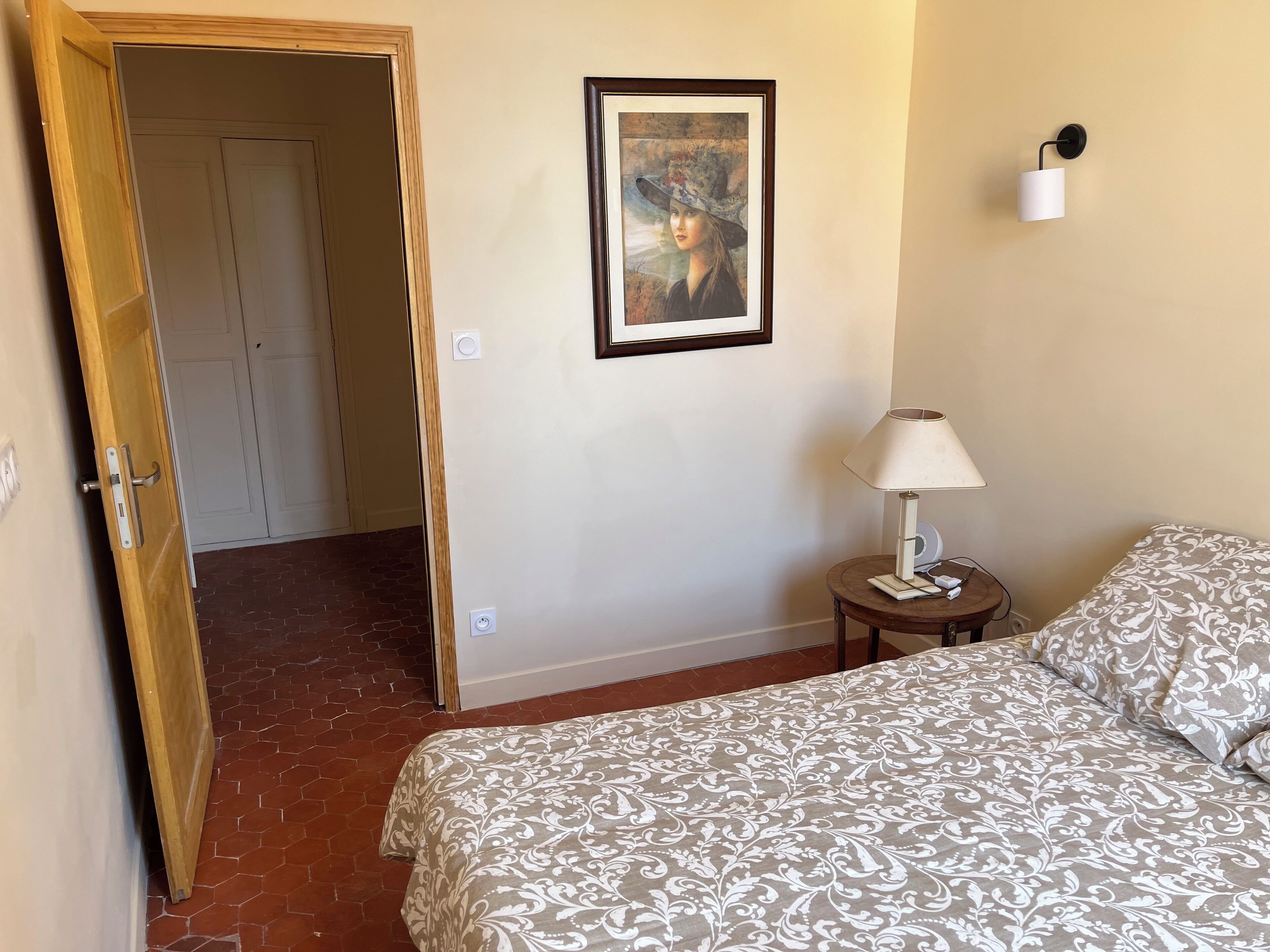A hotel room with a bed, a nightstand, a lamp, a framed picture on the wall, and a door leading to another room.