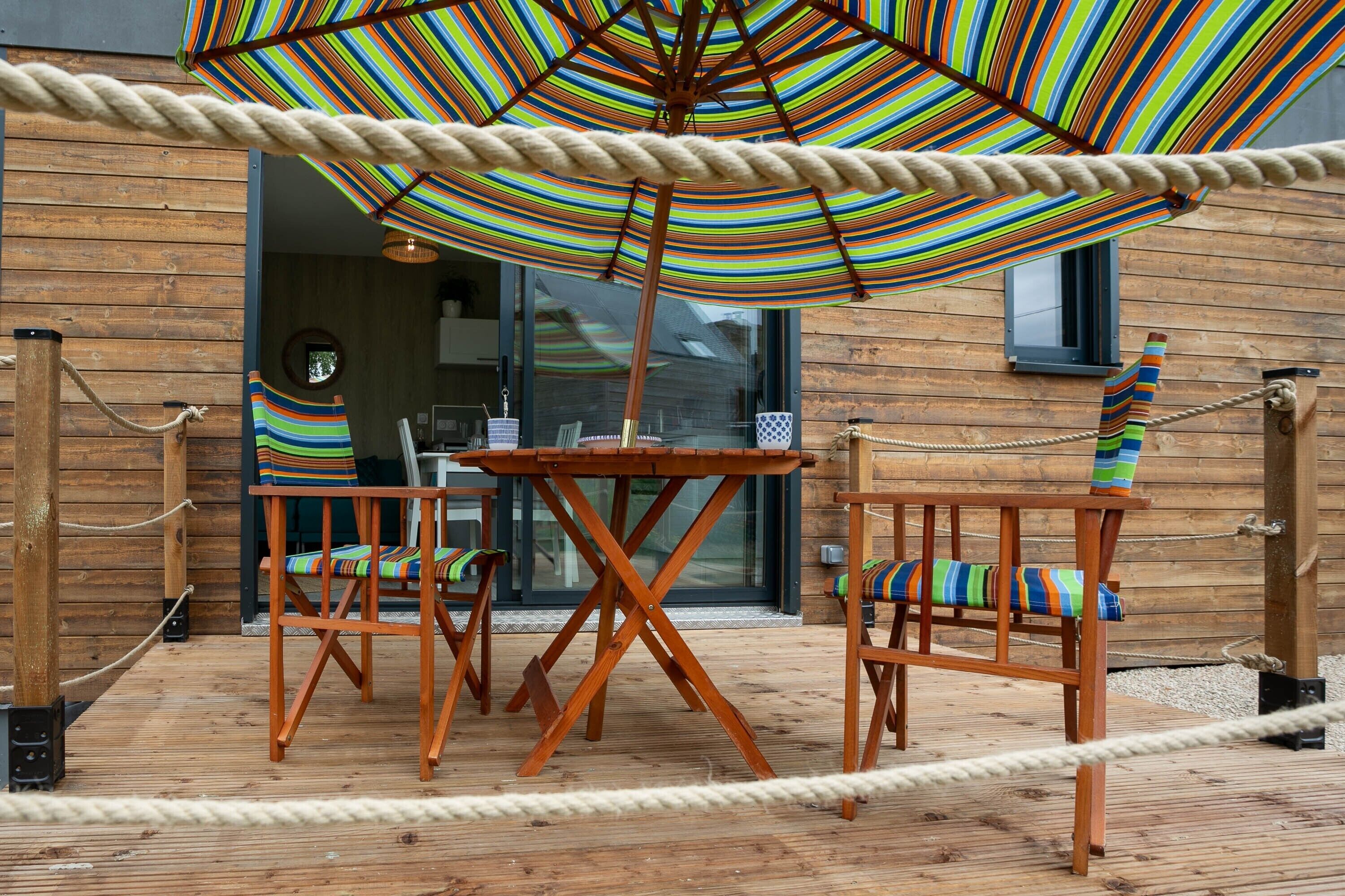 Une terrasse en bois avec une barrière en corde, un parasol à rayures et deux chaises en bois dotées de coussins colorés.