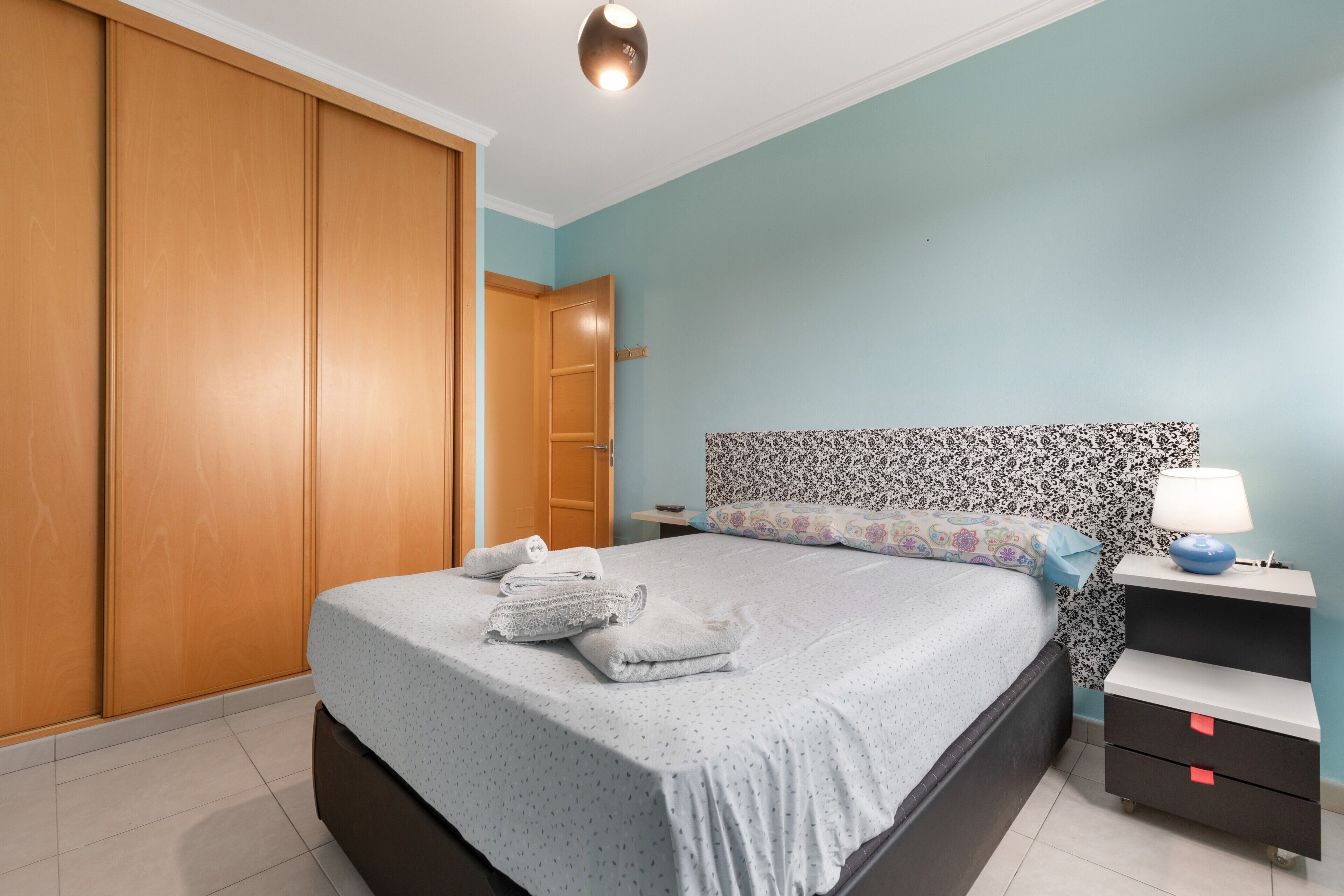 A bedroom with a bed, a nightstand, a lamp, a wardrobe, and a door.