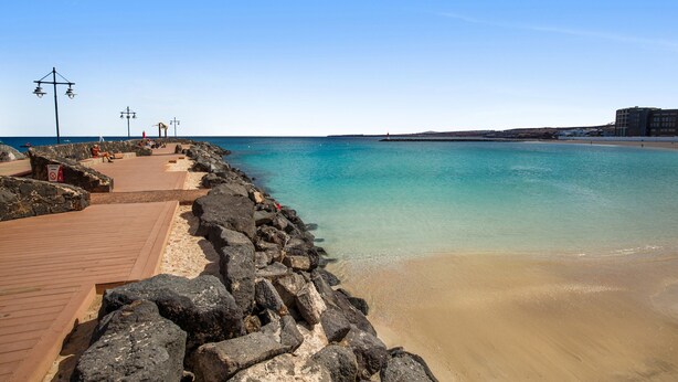 Una playa con un camino de madera, costa rocosa y un edificio al fondo.