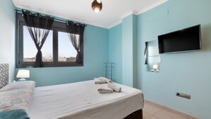 Un dormitorio con cama, ventana con cortinas, televisor montado y lámpara de noche.