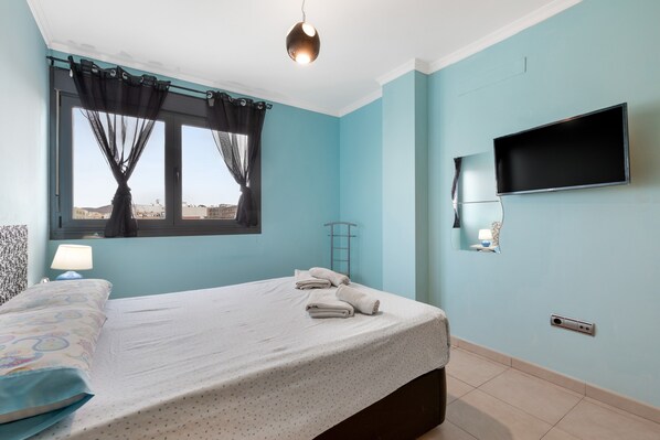 Un dormitorio con cama, ventana con cortinas, televisor montado y lámpara de noche.