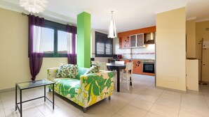 Una sala de estar con sofá, una mesa de centro de vidrio, una mesa de comedor con sillas, una zona de cocina y un ventanal con cortinas.
