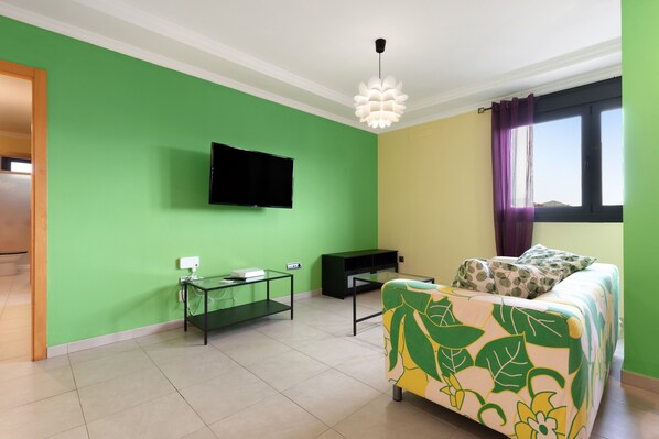 A room with a green wall, a flat-screen TV mounted above a glass table, a bed with a floral bedspread, and a window with purple curtains.
